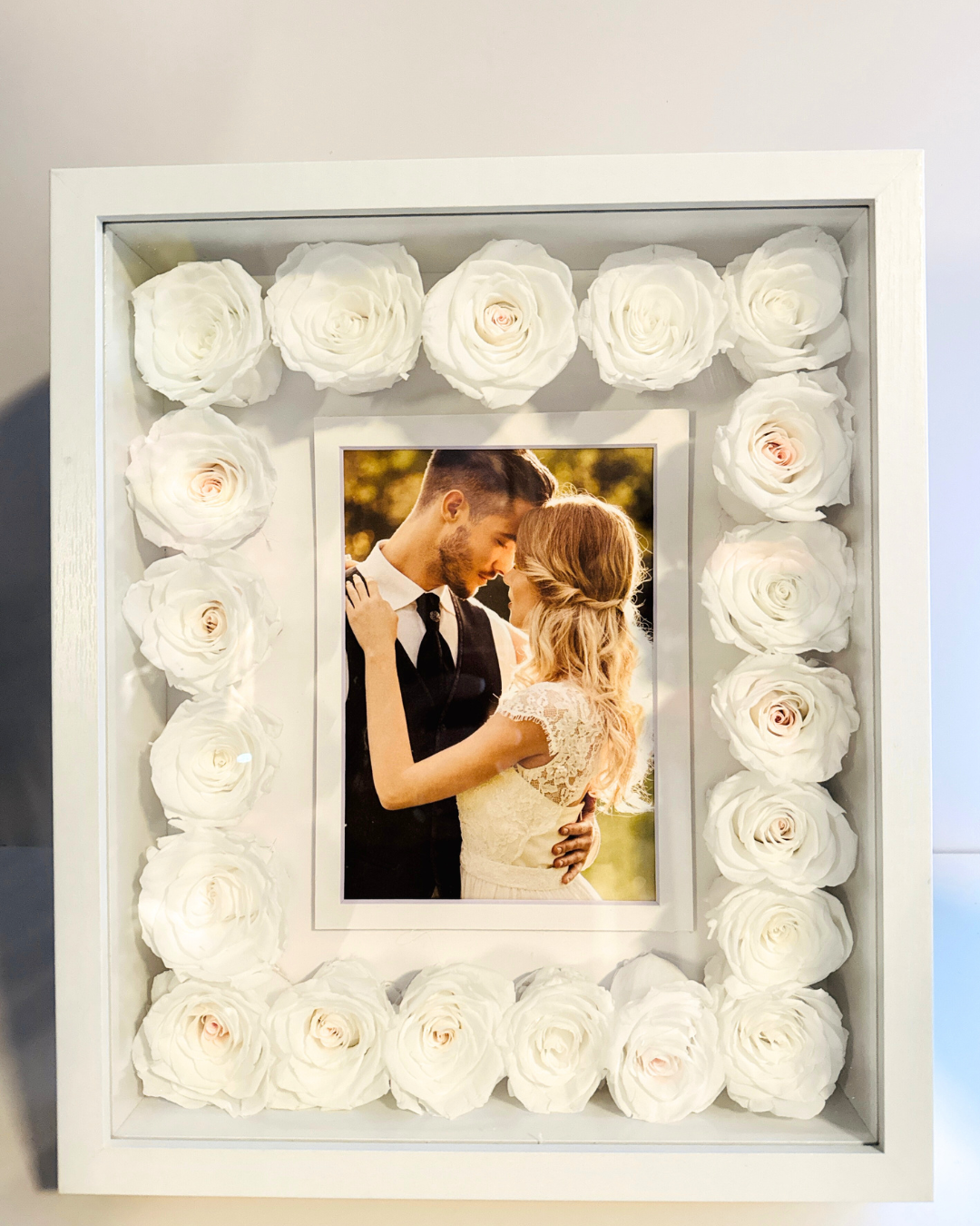 A white picture frame shadow box with preserved roses arranged around the border, containing a photo of a couple embracing | Fleur Reverie