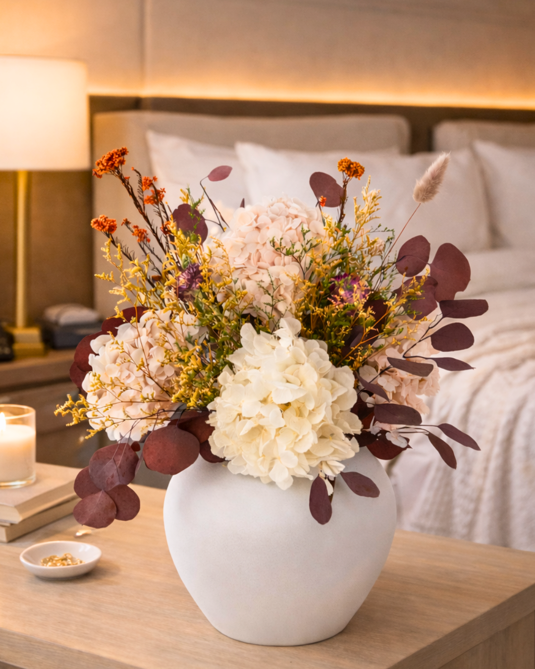 Preserved Floral arrangement in a white vase on a nightstand with a bed in the background | Fleur Reverie