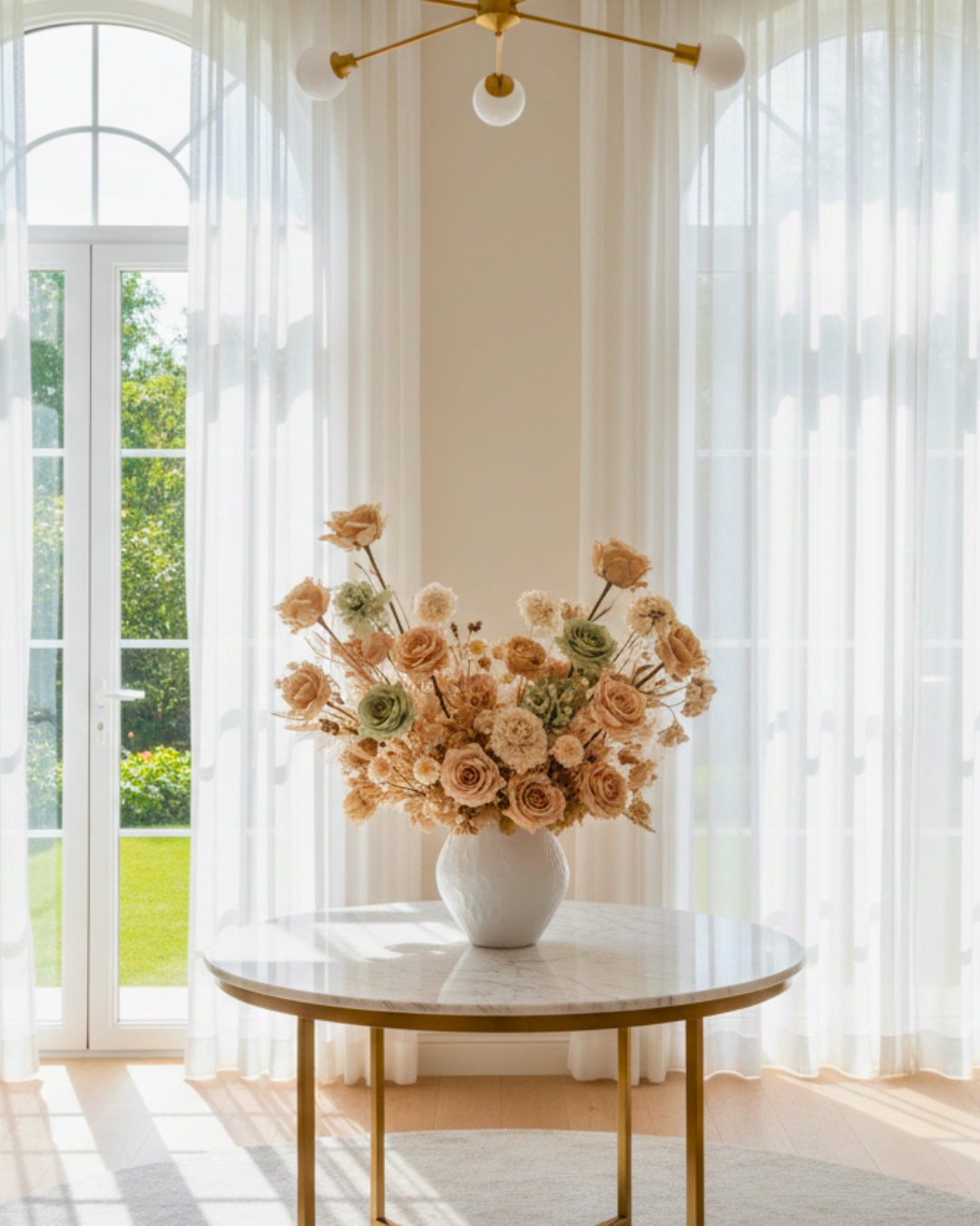 Decorative preserved floral arrangement in a white vase on a marble table with sheer curtains and a chandelier in the background. | Fleur Reverie