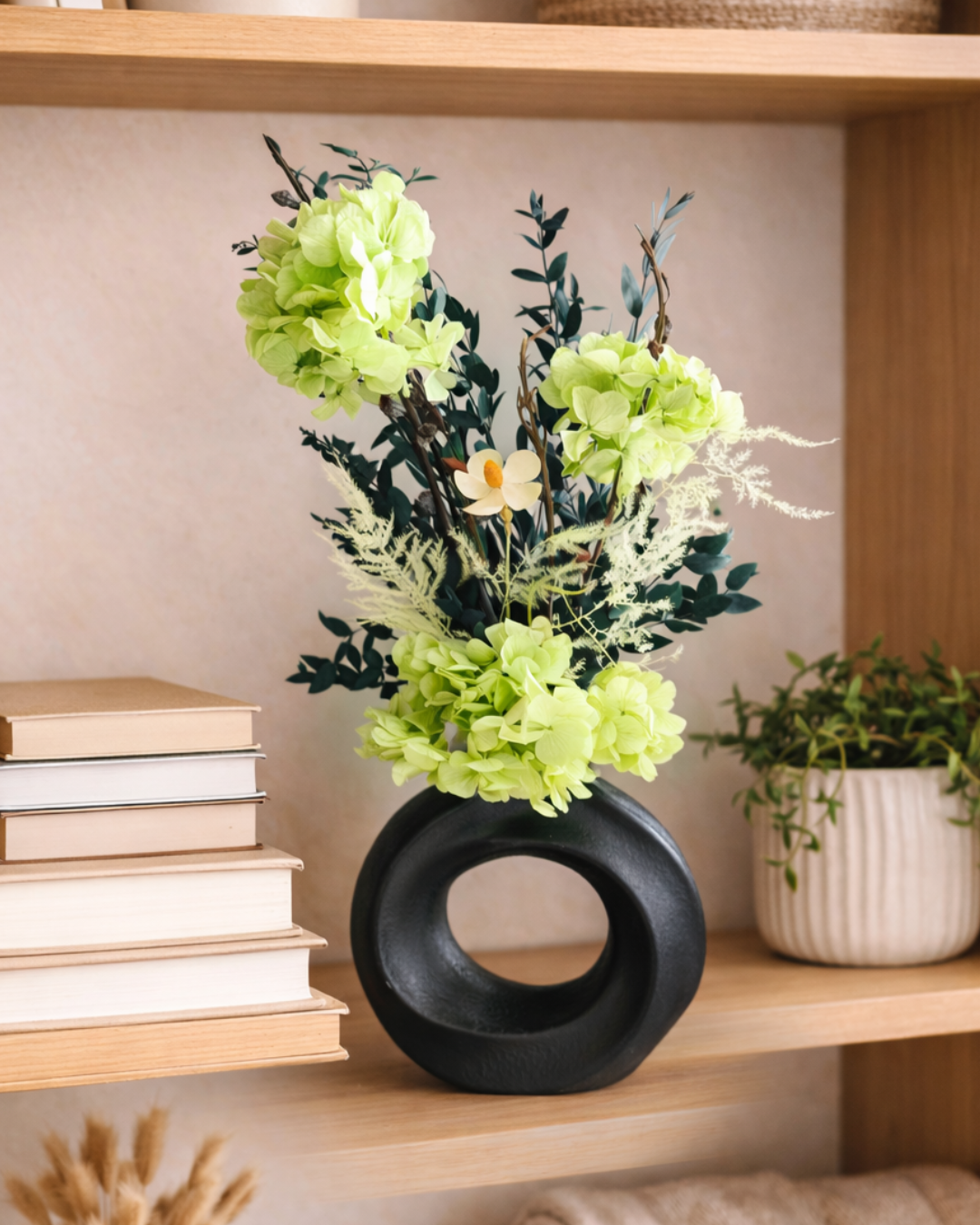 Black vase with green preserved flowers on a wooden shelf with books and another plant. | Fleur Reverie