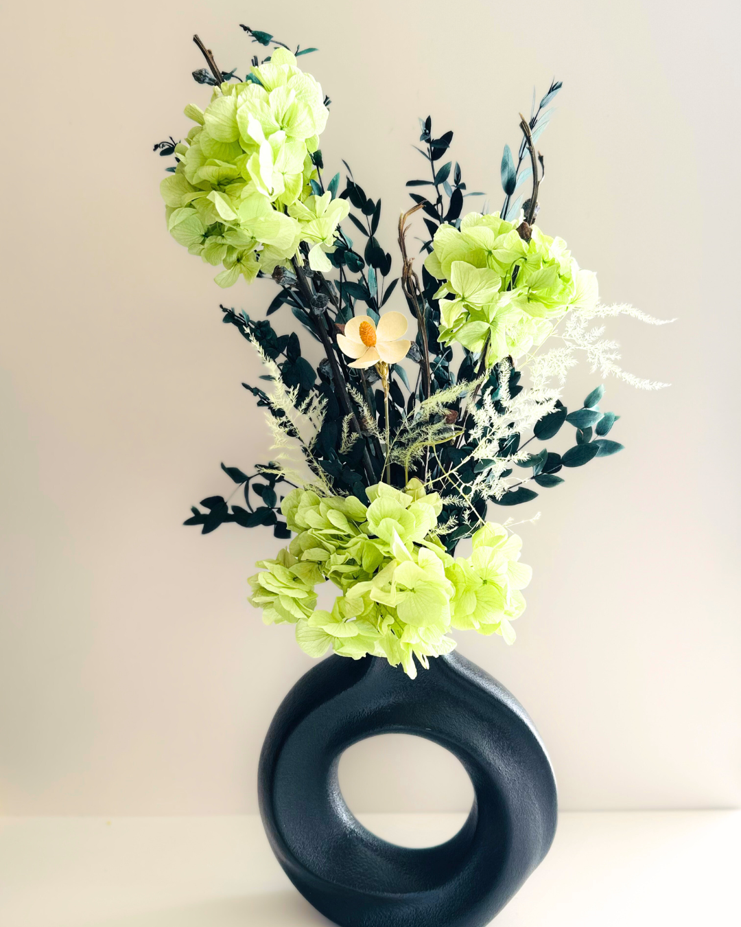 A matte black ceramic donut-shaped vase with preserved large green hydrangeas and decorative foliage inside.