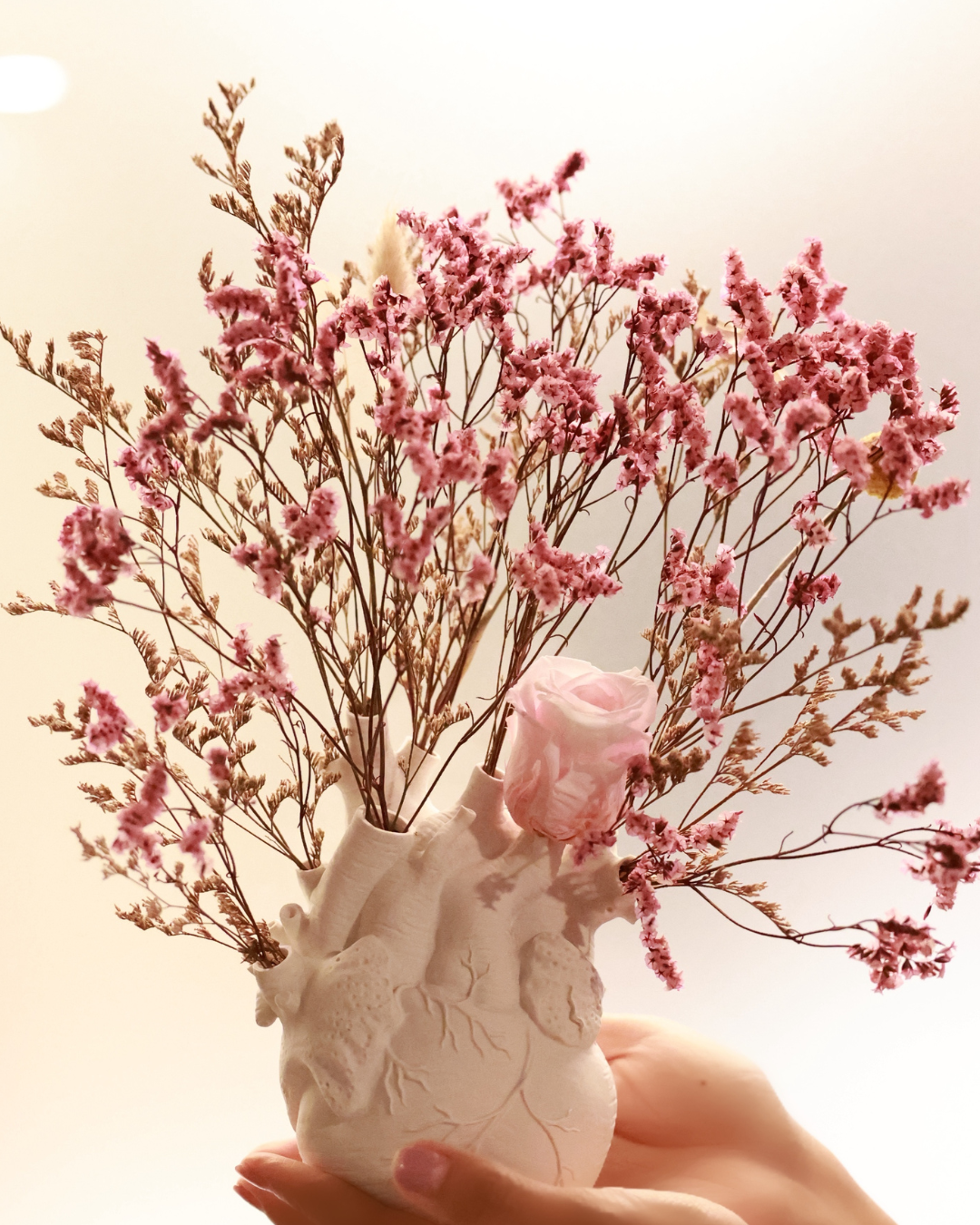 A matte white ceramic vase in the shape of an anatomical heart, filled with preserved blush-toned flowers, held in a person's hands | Fleur Reverie