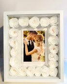 A white picture frame shadow box with preserved roses arranged around the border, containing a photo of a couple embracing | Fleur Reverie