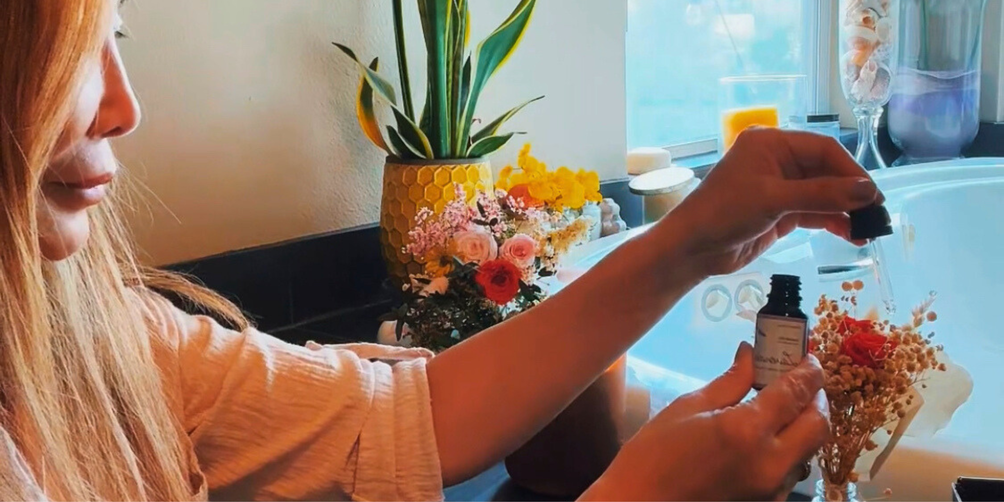 A woman putting essential oil to dried flowers next to a bath tub with vibrant dried flower bouquet on the side