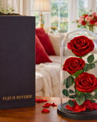 Red roses in a glass dome with 'Fleur Reverie' box on a wooden surface.