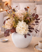 White vase with a bouquet of preserved flowers on a table in a cozy living room setting | Fleur Reverie