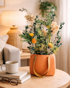 Preserved Floral arrangement in a leather handle bag on a table with a cup and books in a cozy living room.|Fleur Reverie