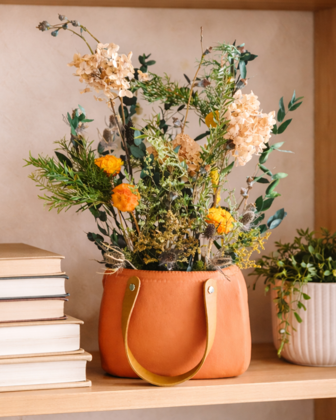 Preserved Floral arrangement in a leather handle bag on a shelf with books and another plant.|Fleur Reverie