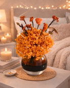 Vase with orange dried flowers on a table next to a lit candle and books, with a cozy room | Fleur Reverie