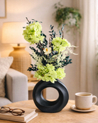 Decorative black vase with green preserved flowers on a table in a living room setting.