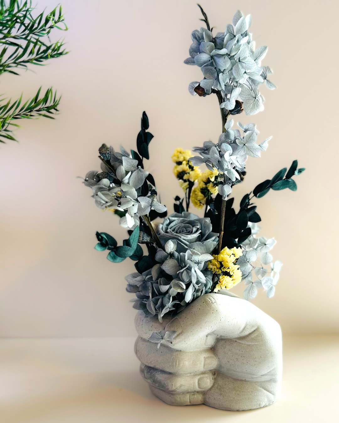 A concrete vase in the shape of a fist holding dried flowers in shades of gray and yellow, placed on a surface with a plant in the background | Fleur Reverie