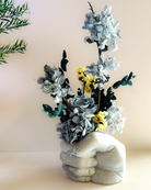 A concrete vase in the shape of a fist holding dried flowers in shades of gray and yellow, placed on a surface with a plant in the background | Fleur Reverie