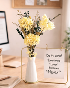 Decorative setup with preserved flowers in a metal vase and a motivational sign on a desk.