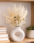 White vase with dried flowers on a shelf with books and another plant.